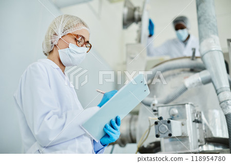 Side view of female technician in protective mask taking notes on notepad while inspecting manufacturing machine on shop floor of pharmaceutical factory, copy space 118954780