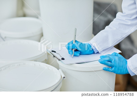 Cropped shot of unrecognizable production worker in blue vinyl gloves taking notes in notepad controlling processes at pharmaceutical manufacturing plant while writing on plastic container, copy space 118954788