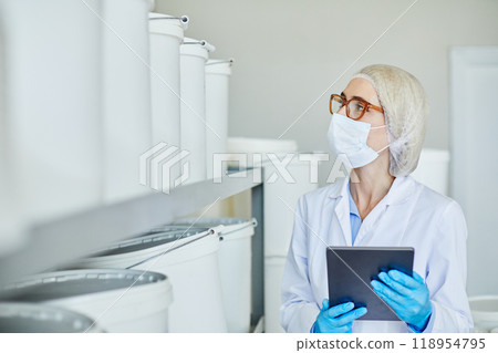 Female process technician with digital tablet in hands taking inventory in stockroom of pharmaceutical factory inspecting large plastic containers filled with pills, copy space 118954795