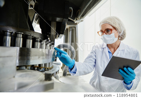 Professional female process technician in vinyl gloves and mask inspecting machinery while pointing at punch tools of tablet press machine at pharmaceutical manufacturing plant 118954801