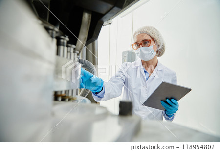 Shot from pill press machine of female process technician in sterile classroom clothing holding digital tablet while inspecting manufacturing equipment at pharmaceutical factory, copy space 118954802