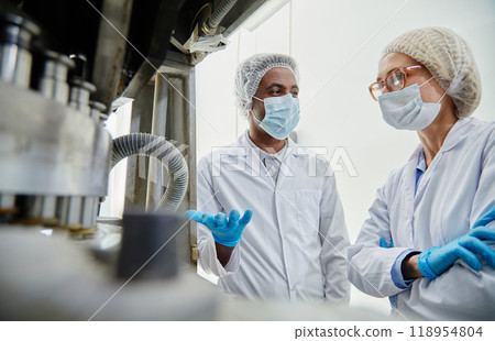 Shot from tablet manufacturing machine of male process technician in white coat inspecting equipment talking to female coworker at pharmaceutical factory 118954804