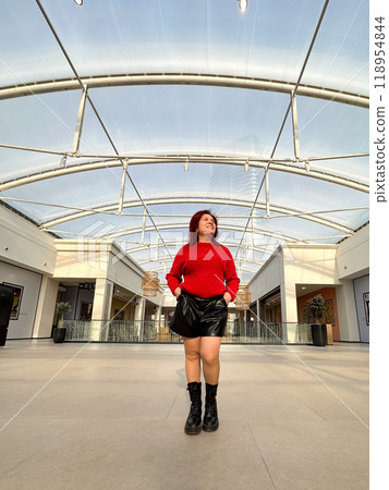 Millennial woman poses confidently in an open, modern environment, wearing a vibrant red sweater and black boots. Joyful expression captures a sense of freedom and individuality 118954844