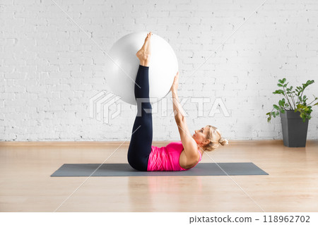 Straight leg hip crunch exercise with fitness ball. Fit Caucasian woman in red tank top does workout with props in loft studio indoor. 118962702