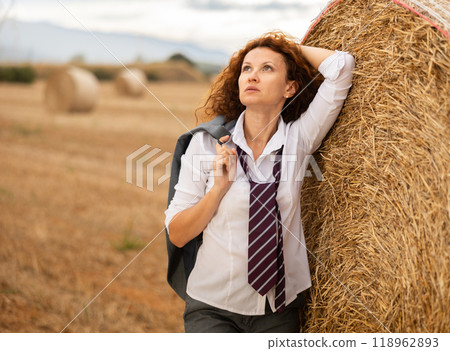 Young woman in white blouse and gray business suit standing on soft plaid mat near stack of straw 118962893