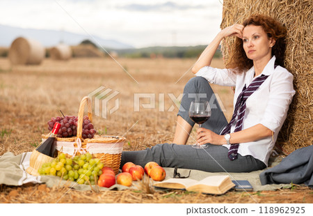 Calm businesswoman sitting by hay bale with wine and book 118962925