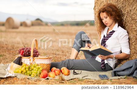 Woman in white blouse and suit spends time in field, settled down in nature with book and drink Woman in white blouse and suit spends time in field, settled down in nature with book and drink 118962964