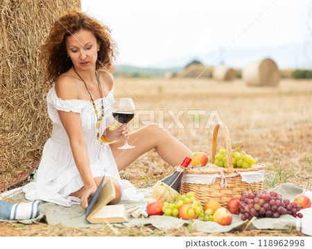 Carefree woman in white sitting on picnic near haystack with wine and book 118962998