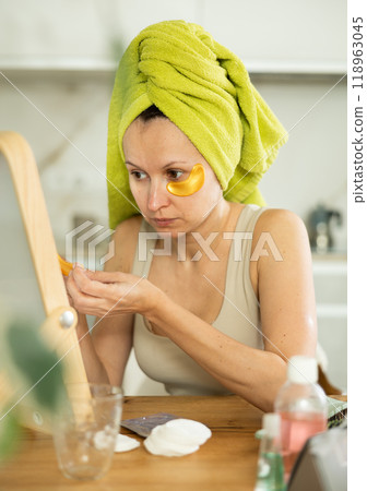 Middle-aged woman applying eye patch mask on her face sitting in front of the mirror Middle-aged woman applying eye patch mask on her face sitting in front of the mirror 118963045