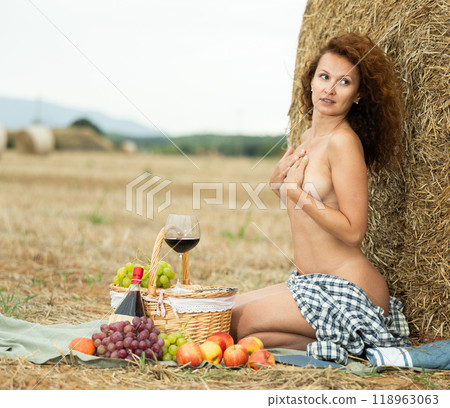 Topless woman in gray checkered unbuttoned shirt holds glass of wine, sitting next to straw pile Topless woman in gray checkered unbuttoned shirt holds glass of wine, sitting next to straw pile 118963063