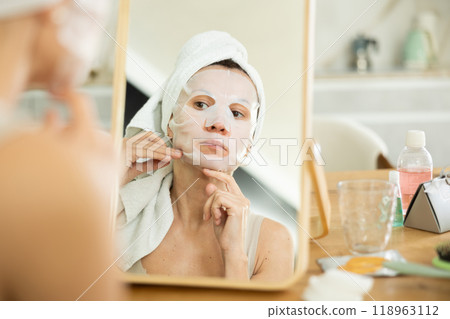 Middle-aged woman applying tissue mask on her facial skin sitting in front of the mirror Middle-aged woman applying tissue mask on her facial skin sitting in front of the mirror 118963112