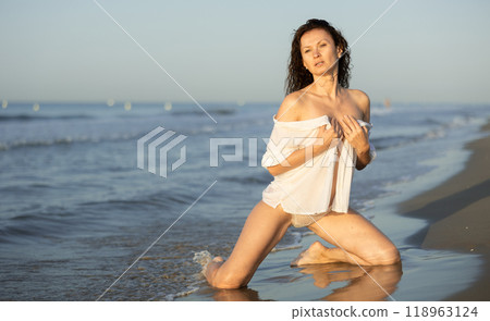 Portrait of female posing in white lingerie and mesh cape on knees on beach near sea at sunset 118963124