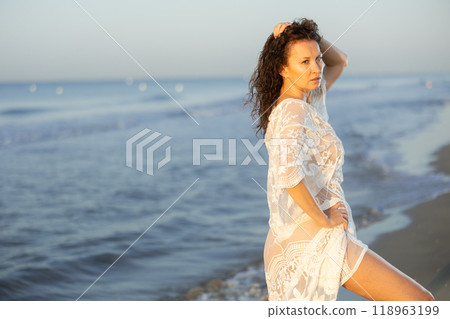 Sexy woman in white lingerie and mesh cape walking along the seashore in surf at sunset 118963199
