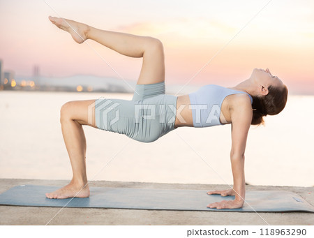 Sporty energetic experienced young woman yoga trainer performing upward plank pose on rug on beach at sunrise. Fitness concept Sporty energetic experienced young woman yoga trainer performing upward plank pose on rug on beach at sunrise. Fitness concept 118963290