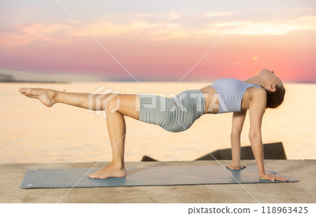Sporty energetic experienced young woman yoga trainer performing upward plank pose on rug on beach at sunrise. Fitness concept 118963425