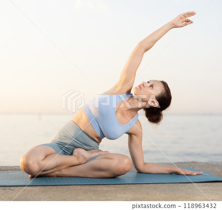 Near sea during sunset, girl performs variations of padmasana with body tilt 118963432