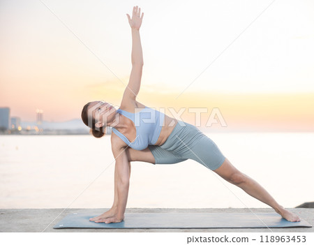 Skilled happy young female teacher demonstrating variation of warrior pose of yoga on seashore at sunrise 118963453