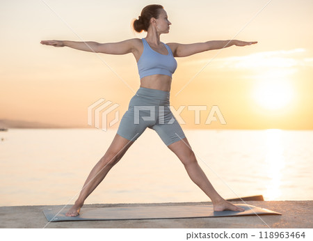 Near sea during sunset, girl performs variations of virabhadrasana 118963464
