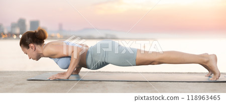 Near sea during sunset, girl performs Chaturanga dandasana 118963465