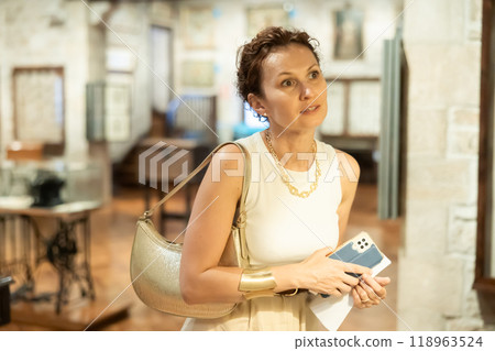 Portrait of middle-aged woman with smartphone in her hands looking at an exhibition in historical museum Portrait of middle-aged woman with smartphone in her hands looking at an exhibition in historical museum 118963524