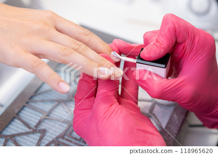 Female manicurist master in pink gloves carefully applies base for gel polish to a clients nails in a salon. 118965620