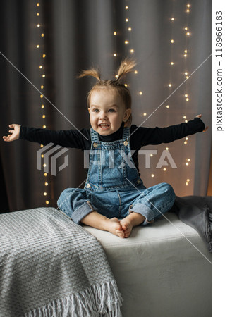 Smiling toddler girl in denim overalls with fun hairstyle indoors 118966183