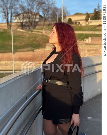Millennial woman stands confidently in sunlight, with vibrant red hair flowing and dressed in a chic black outfit. Background of a clear blue sky and distant scenery enhances the dynamic and modern 118971932