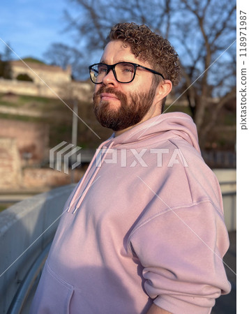 Millennial man enjoys a peaceful moment autumn outdoors, wearing a soft pink hoodie and glasses. Background of trees and historic architecture creates an atmosphere of tranquility and contemplation 118971987