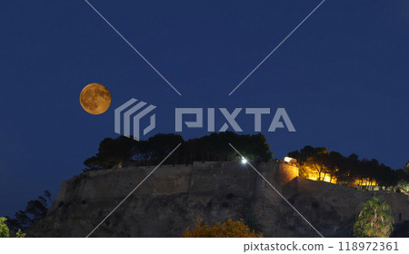 A majestic full moon rises over an ancient castle, casting a mystical glow on its towering walls and surrounding trees at night, Denia, Spain 118972361