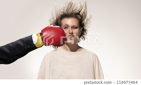 Red boxing glove makes contact with woman's cheek, her hair wild as she reacts to sudden impact against white studio background. 118973464