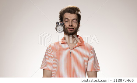 Alarm clock smashes into man's cheek, his face squished from force against white studio background. Time management. 118973466