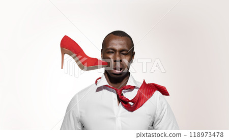 Bright red high heel shoe crashes into African-American man's face, distorting his expression against white studio background. 118973478