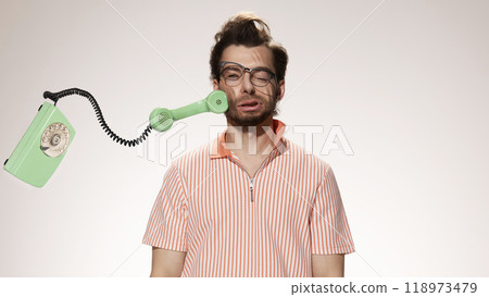 Rotary phone handset slams into man's cheek, his scrunched face reflecting impact against white studio background. Concept of frustration. 118973479