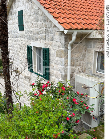 Charming stone house with red tile roof and blooming red flowers surrounding the window. Picturesque example of traditional architecture blending with nature in a quaint setting Charming stone house with red tile roof and blooming red flowers surrounding the window. Picturesque example of traditional architecture blending with nature in a quaint setting 118975989