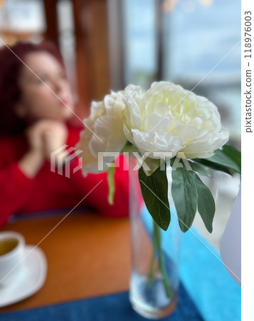 Focus on a delicate white flower in a vase with a blurred woman in background, dressed in red, depicting a serene indoor scene. Contrasting colors create a sense of tranquility and contemplation. Focus on a delicate white flower in a vase with a blurred woman in background, dressed in red, depicting a serene indoor scene. Contrasting colors create a sense of tranquility and contemplation. 118976003