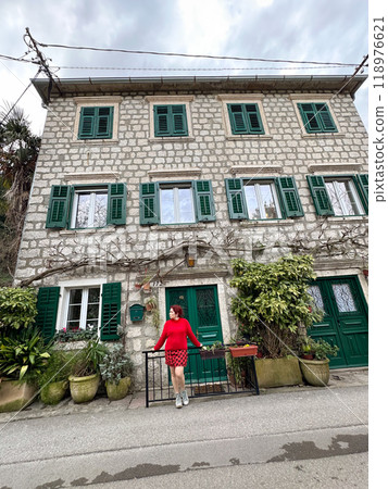 Charming stone building with green shutters and millennial woman in red standing in front. a picturesque scene capturing the essence of rustic architecture and fashion. Travel and journey concept Charming stone building with green shutters and millennial woman in red standing in front. a picturesque scene capturing the essence of rustic architecture and fashion. Travel and journey concept 118976621