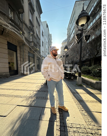 Millennial man stands confidently on a sunlit city street, wearing a cozy jacket and stylish hat. Urban landscape and soft morning light create a scene of casual elegance and modern lifestyle 118976678