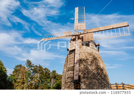 Old stone windmill stands against a clear blue sky with gentle clouds. 118977935