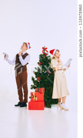 Couple wearing Santa hats, reviewing Christmas lists with pens in hand, standing near decorated tree and holiday presents, planning their holiday. 118978960