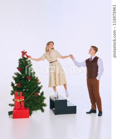 Woman stands on step, playfully reaching for Christmas tree while holding hands with beloved man, who helps her balance. 118978961