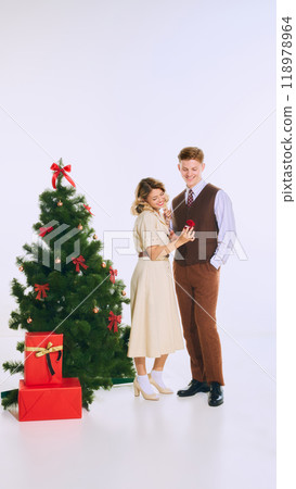 Woman shows man small red gift box, standing by Christmas tree, surrounded by holiday cheer, as they share loving moment. Woman shows man small red gift box, standing by Christmas tree, surrounded by holiday cheer, as they share loving moment. 118978964