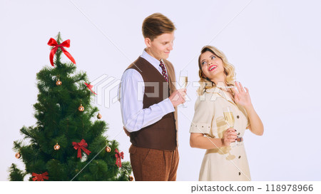 Couple joyfully toasts with champagne beside decorated Christmas tree, dressed in vintage outfits, celebrating festive moment with smiles. 118978966
