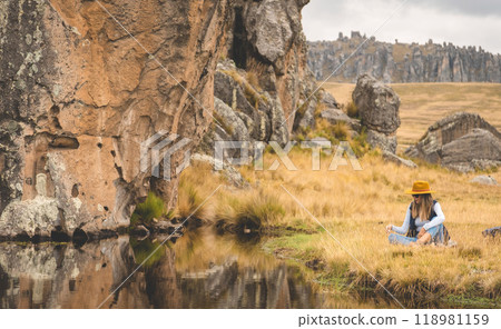 A woman sits in a field next to a river, tourist in the peruvian andes, Huaillay, Pasco, Peru. 118981159