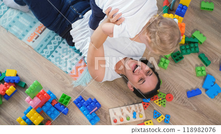Happy mother playfully lifts her baby while lying on the floor, surrounded by bright toys, creating a fun and lively scene 118982076