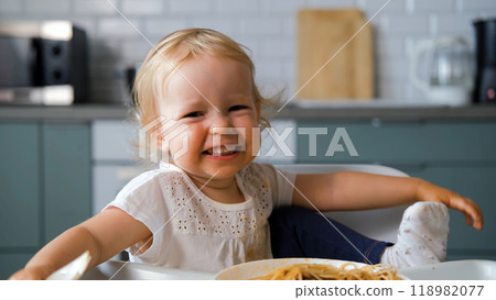 Cheerful baby in a high chair attempts to eat delicious homemade pasta bolognese with a fork in the kitchen 118982077