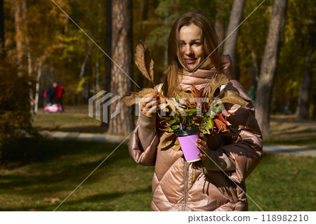 Smiling woman in a pink jacket with a glass of coffee tea in autumn park Smiling woman in a pink jacket with a glass of coffee tea in autumn park 118982210