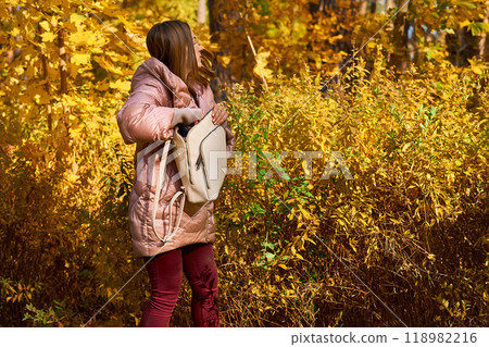 Pretty woman in pink jacket and red jeans takes something out of bag in autumn Pretty woman in pink jacket and red jeans takes something out of bag in autumn 118982216