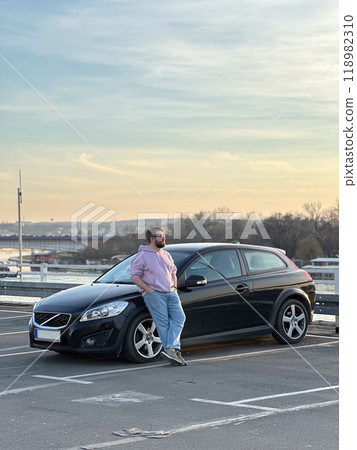 Millennial man in a casual hoodie leans confidently against a sleek black car in a parking lot. Peaceful evening sky forms a serene backdrop to this moment of relaxation and contemplation Millennial man in a casual hoodie leans confidently against a sleek black car in a parking lot. Peaceful evening sky forms a serene backdrop to this moment of relaxation and contemplation 118982310