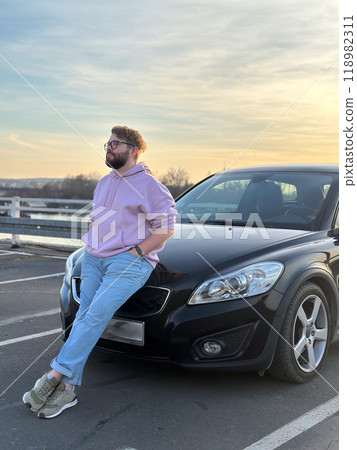 Millennial man in a casual hoodie leans confidently against a sleek black car in a parking lot. Peaceful evening sky forms a serene backdrop to this moment of relaxation and contemplation Millennial man in a casual hoodie leans confidently against a sleek black car in a parking lot. Peaceful evening sky forms a serene backdrop to this moment of relaxation and contemplation 118982311