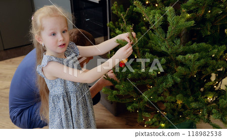 Young girl delicately hanging a christmas ornament on a tree branch with her father's assistance. Little girl decorating Christmas tree with vintage toys 118982538